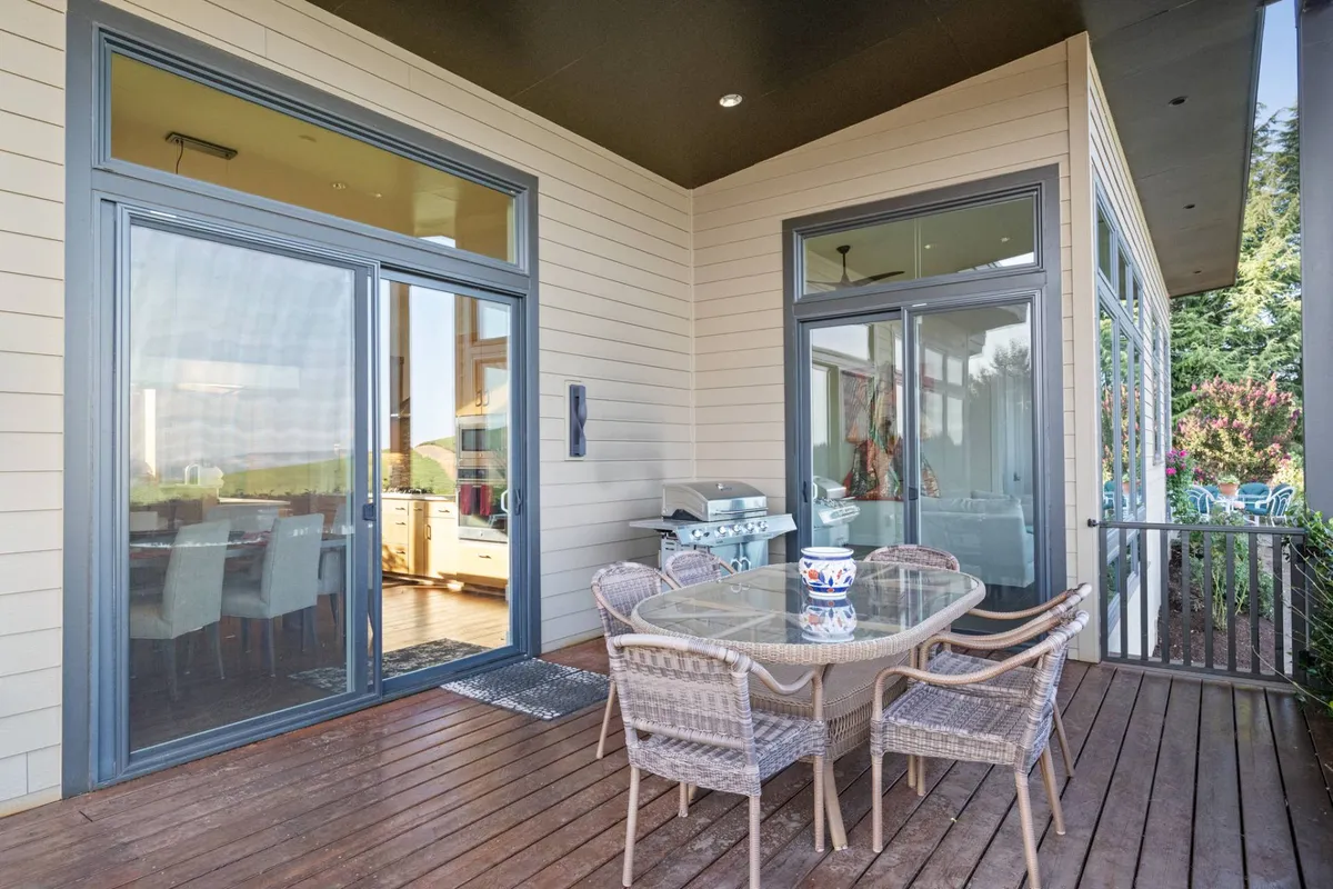 An outdoor dining space, complete with a table, grill, and glass doors leading inside, is surrounded by greenery. The area invites relaxation and social gatherings, suggesting evenings spent dining outside in a picturesque natural setting.