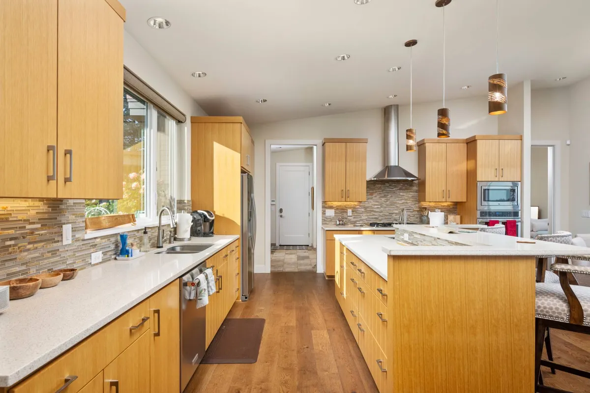 A spacious, light-filled kitchen featuring a generous kitchen island, stainless steel professional-grade appliances, rich wood cabinetry, and ample natural light streaming in from large windows. The open design encourages both meal preparation and socializing, with plenty of workspace visible.