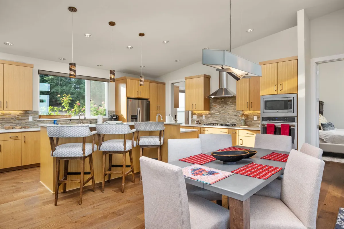 A kitchen-dining area with a kitchen island, bar stools for casual seating, red-accented dining table, and large windows providing sunlight and a hint of the outdoor landscape. The scene exudes warmth and a sense of gathering, ideal for both casual bites and group celebrations.