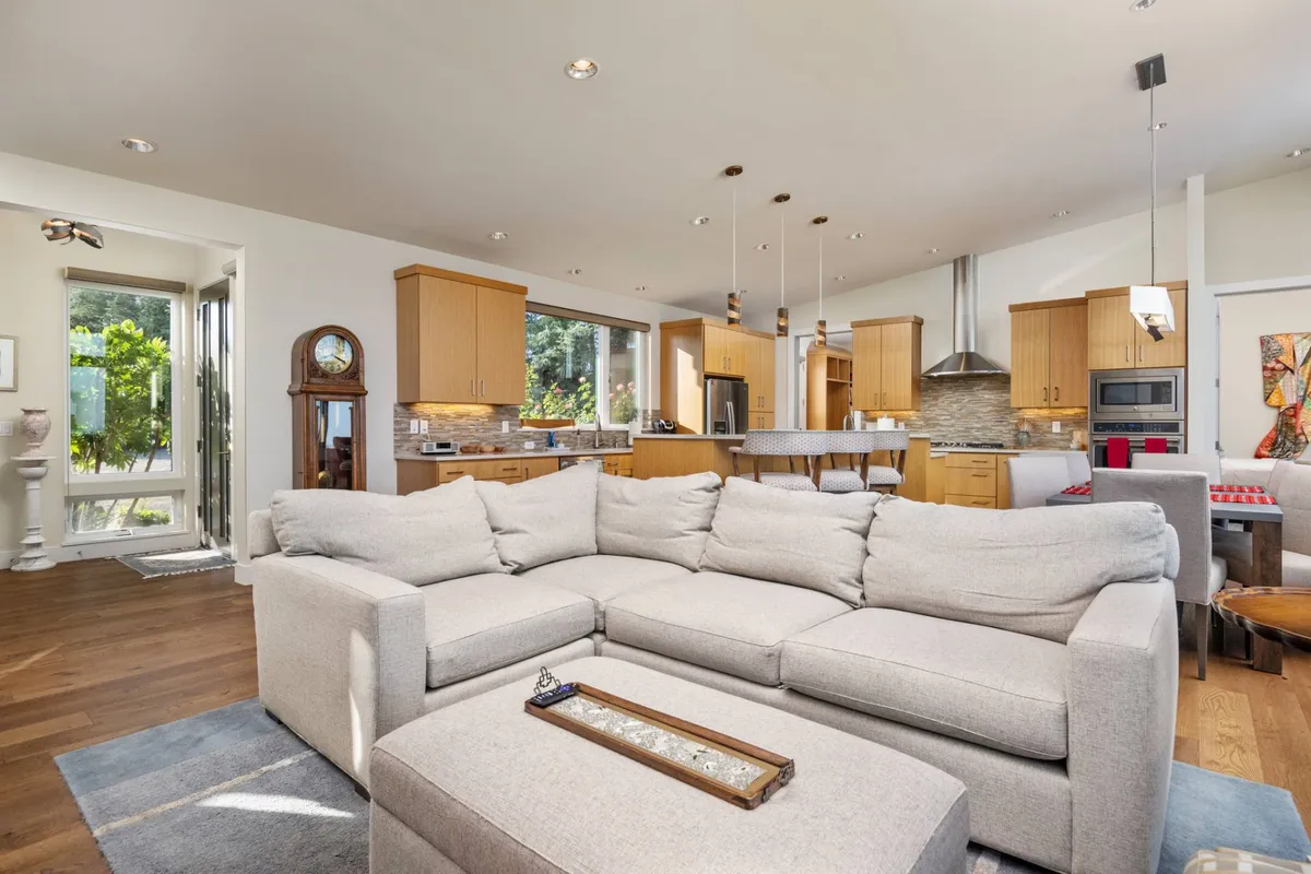 This image shows the open-plan living and dining area with a large sectional sofa, kitchen area, dining table, a clock, and artwork on the wall. The room is filled with natural light, and the arrangement invites social gatherings and cozy family moments.