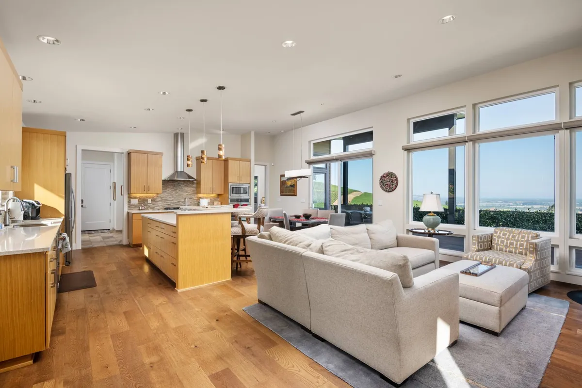 This image highlights a welcoming living area adjacent to an open kitchen and dining table, with large windows showcasing the landscape. The arrangement balances modern comfort with views, inviting guests to relax, cook, and dine while appreciating the beauty beyond.