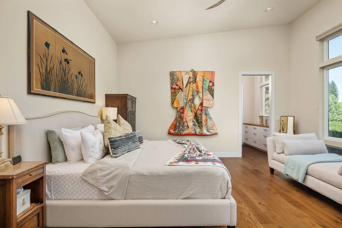 A serene bedroom featuring a comfortable bed with textile art above, a nightstand with a lamp, and a cozy seating area by the window. Soft natural light streams in, creating an inviting space ideal for relaxation and restful mornings.