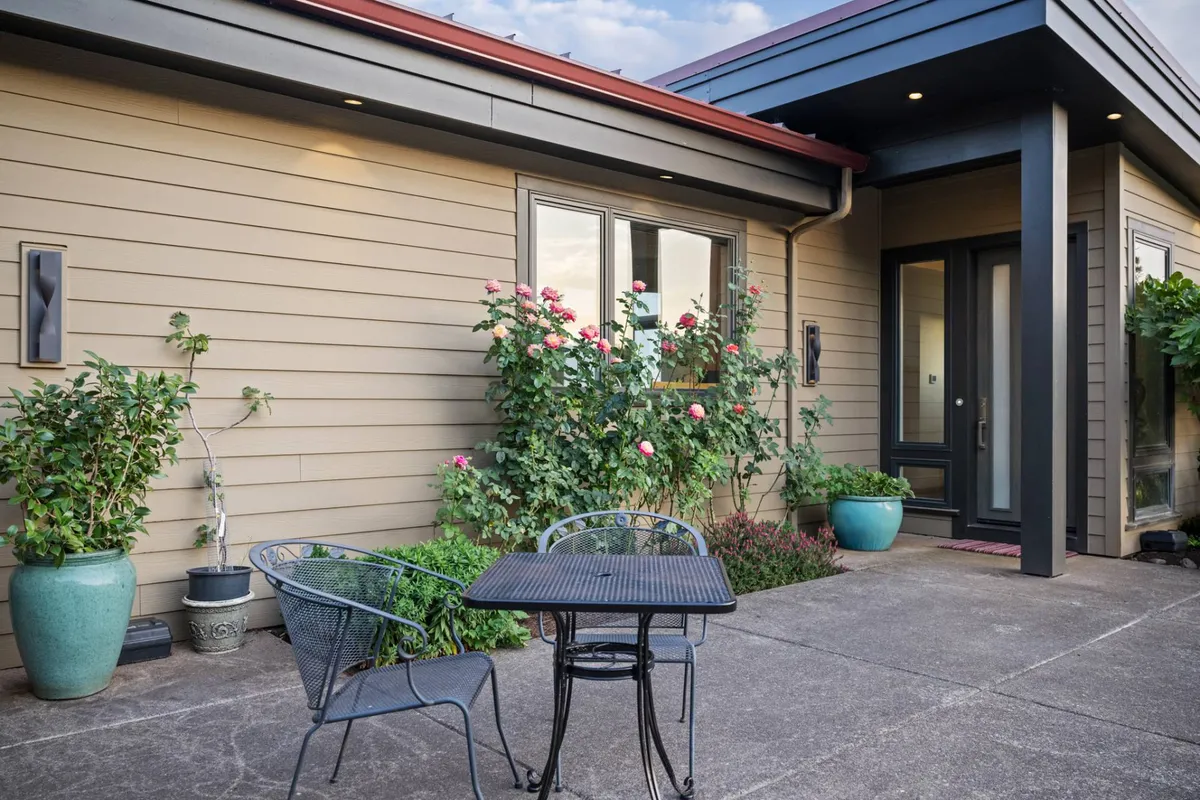The photo features an inviting patio setup with a table and chairs surrounded by flowering plants, just outside the entrance door. The soft morning or evening light enhances the cozy and intimate atmosphere, hinting at peaceful mornings with coffee or romantic evenings under the stars.