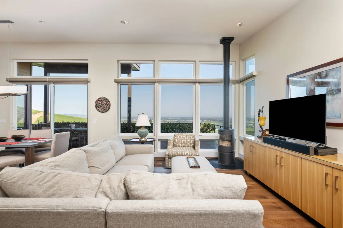 This image captures a spacious great room featuring a large sectional sofa, floor-to-ceiling windows that flood the space with natural light, a wooden media console, a decorative lamp, and a cozy stove. The panoramic windows provide expansive views of the valley, emphasizing the connection between the interior and the stunning landscape beyond.