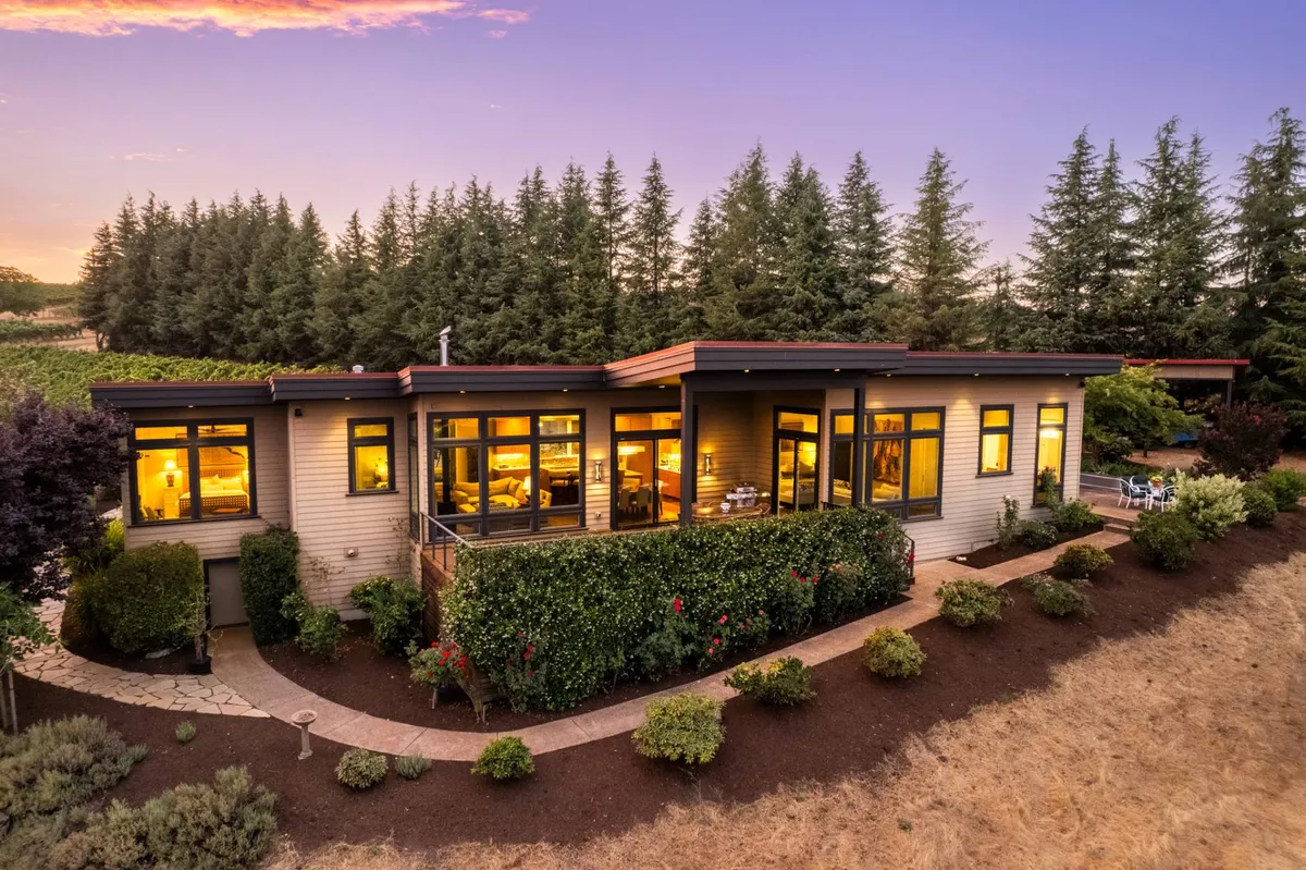 A view of the house enveloped by lush greenery and garden blooms, with a welcoming pathway leading to the entrance. The scene highlights the vibrant landscaping and year-round floral interest of the grounds.