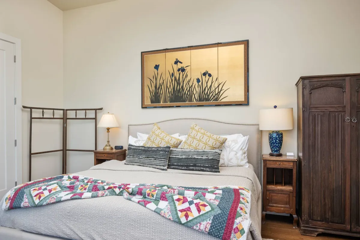 A cozy bedroom with a bed topped by a colorful quilt, framed artwork above, and inviting nightstands with lamps on either side. The ambiance is warm and serene, perfect for starting or ending the day in comfort.