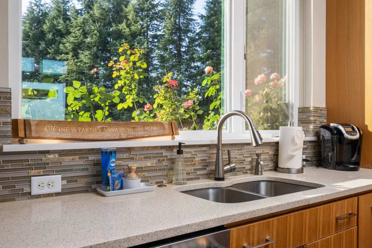 A close-up of a kitchen sink below a window with a view of vibrant flowers outside, accompanied by a wooden sign. The image evokes the feeling of cooking with fresh, garden-picked ingredients just steps away.