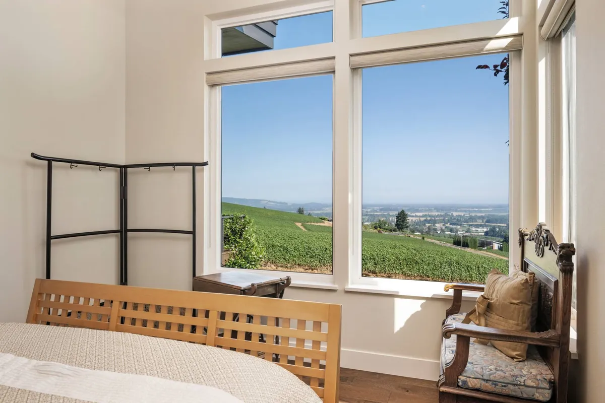 A bedroom with a wooden bed frame, large windows framing vineyard views, an antique chair, and a small table—designed for morning coffee or quiet contemplation. The setting is calm and reflective.